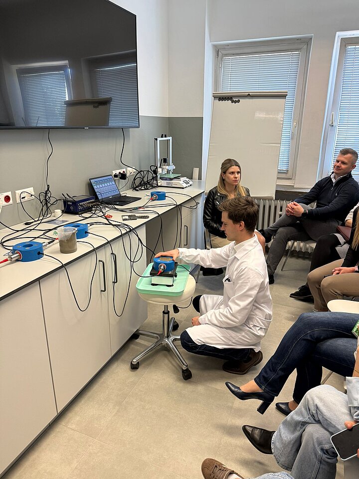 grupa osób siedzi na krzesłach w laboratorium i słuchają mężczyznę ubranego w fartuch laboratoryjny 