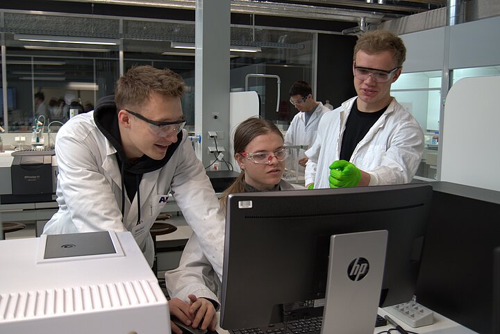 trzy młode osoby w fartuchach laboratoryjnych patrzą na monitor 