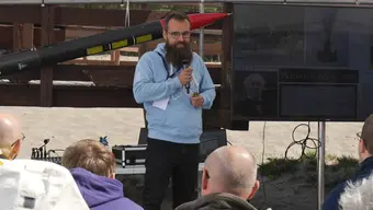 mężczyzna z brodą ubrany w niebieską bluzę stoi z mikrofonem i mówi