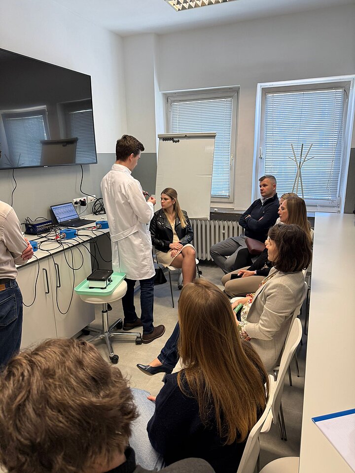 grupa osób siedzi na krzesłach w laboratorium i słuchają mężczyznę ubranego w fartuch laboratoryjny 